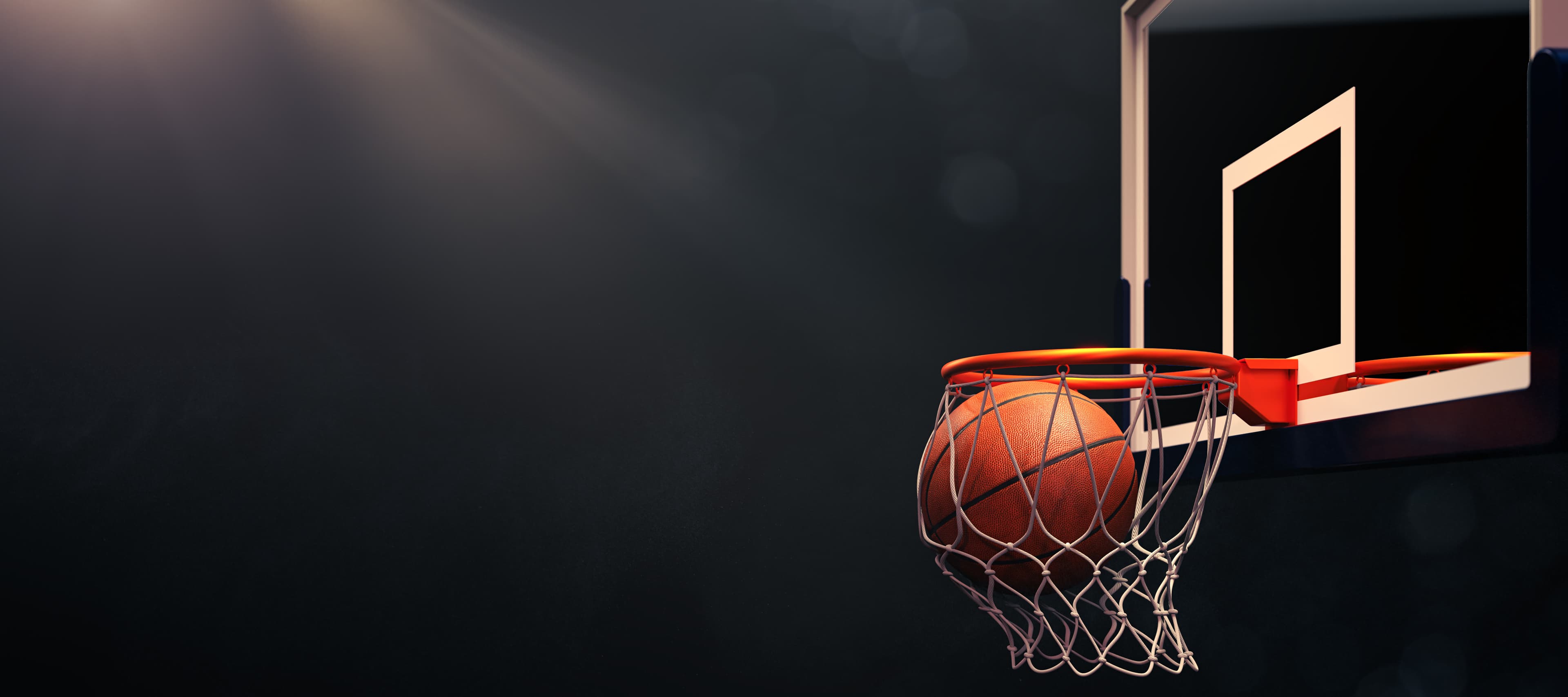A basketball dropping through the net of a lit hoop against a dark court.