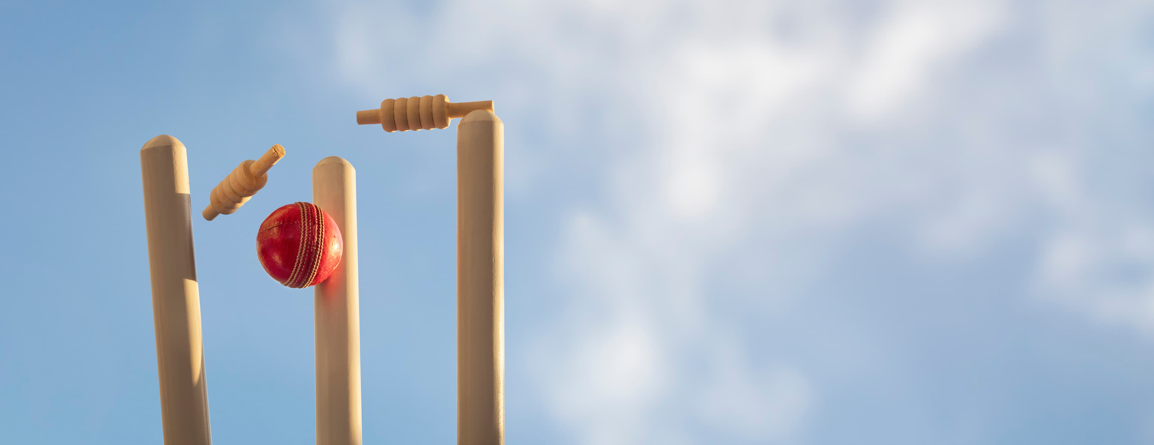 A red cricket ball striking the stumps, bails flying, against a blue sky.
