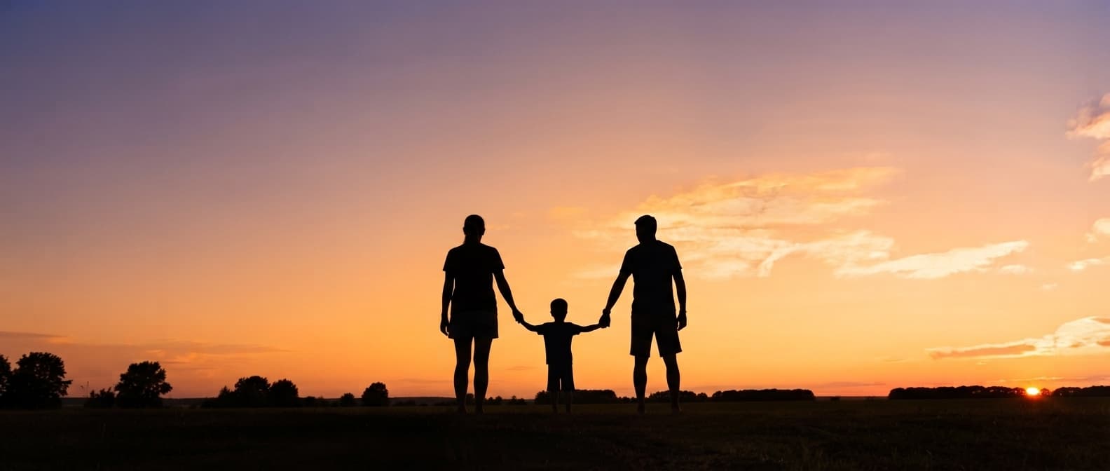 Two adults and a young child stand hand in hand on open grassland, silhouetted against a purple and orange sunset sky.